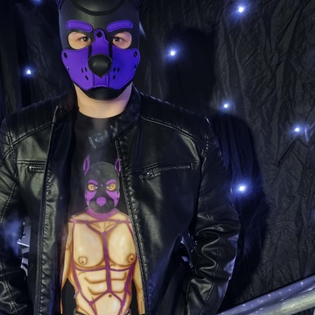 Model wearing black T-shirt, hoodie, and jumper featuring illustrated purple pup hood character from the Alpha Pack series, photographed in a dark studio setting with dramatic lighting, leather jacket styling, and fetish-inspired pup art apparel design.