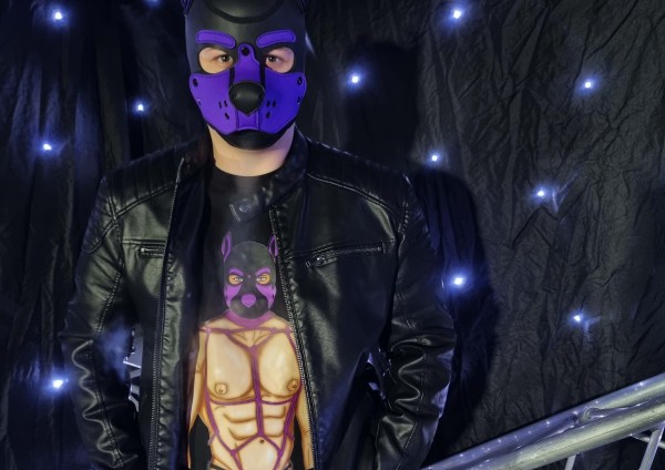 Model wearing black T-shirt, hoodie, and jumper featuring illustrated purple pup hood character from the Alpha Pack series, photographed in a dark studio setting with dramatic lighting, leather jacket styling, and fetish-inspired pup art apparel design.
