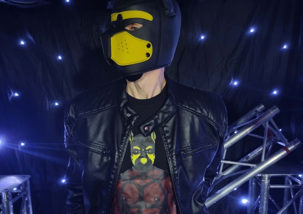 Model wearing black Alpha Pack apparel featuring a yellow pup hood illustration, photographed in a dark studio setting with dramatic lighting and metallic stage structures, showcasing bold fetish-inspired graphic art and confident styling.