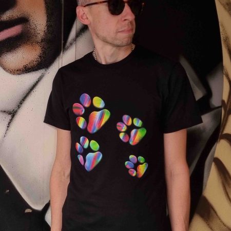 Black t-shirt featuring a vibrant rainbow paw print design displayed on a model in an urban graffiti setting and styled as a flatlay on a dark wood table with accessories. The colourful paw graphic stands out against the deep black fabric, creating a bold yet versatile streetwear look. Also shown in jumper and hoodie variations, highlighting a cohesive fashion collection designed for everyday wear, casual styling, and modern streetwear expression.