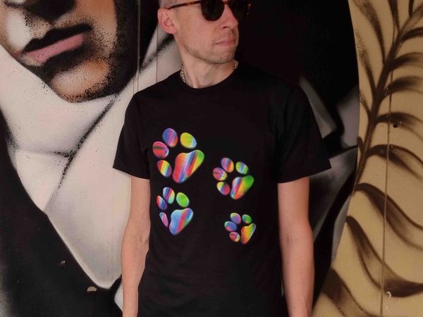 Black t-shirt featuring a vibrant rainbow paw print design displayed on a model in an urban graffiti setting and styled as a flatlay on a dark wood table with accessories. The colourful paw graphic stands out against the deep black fabric, creating a bold yet versatile streetwear look. Also shown in jumper and hoodie variations, highlighting a cohesive fashion collection designed for everyday wear, casual styling, and modern streetwear expression.